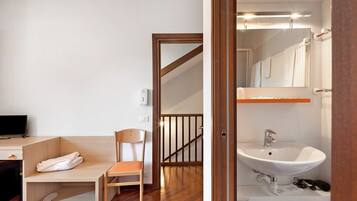 Camera d'albergo con una scrivania in legno, una sedia, un letto e bagno con lavandino e specchio.