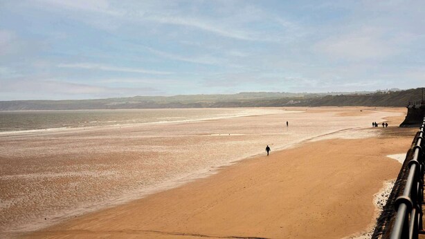A wide sandy beach with a clear sky, a distant hilly landscape, and a few people walking. - Host & Stay | The Beach (Filey)