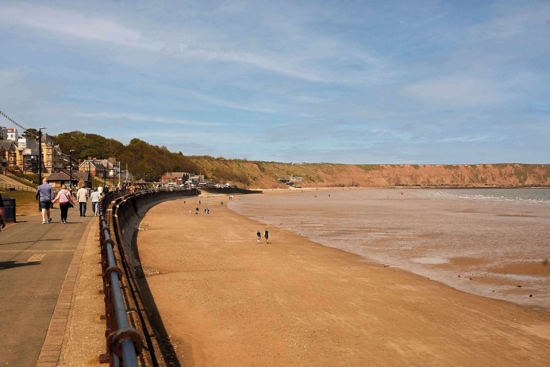 Une promenade en bord de mer avec une plage de sable, une falaise en arrière-plan et des bâtiments qui bordent le chemin.
