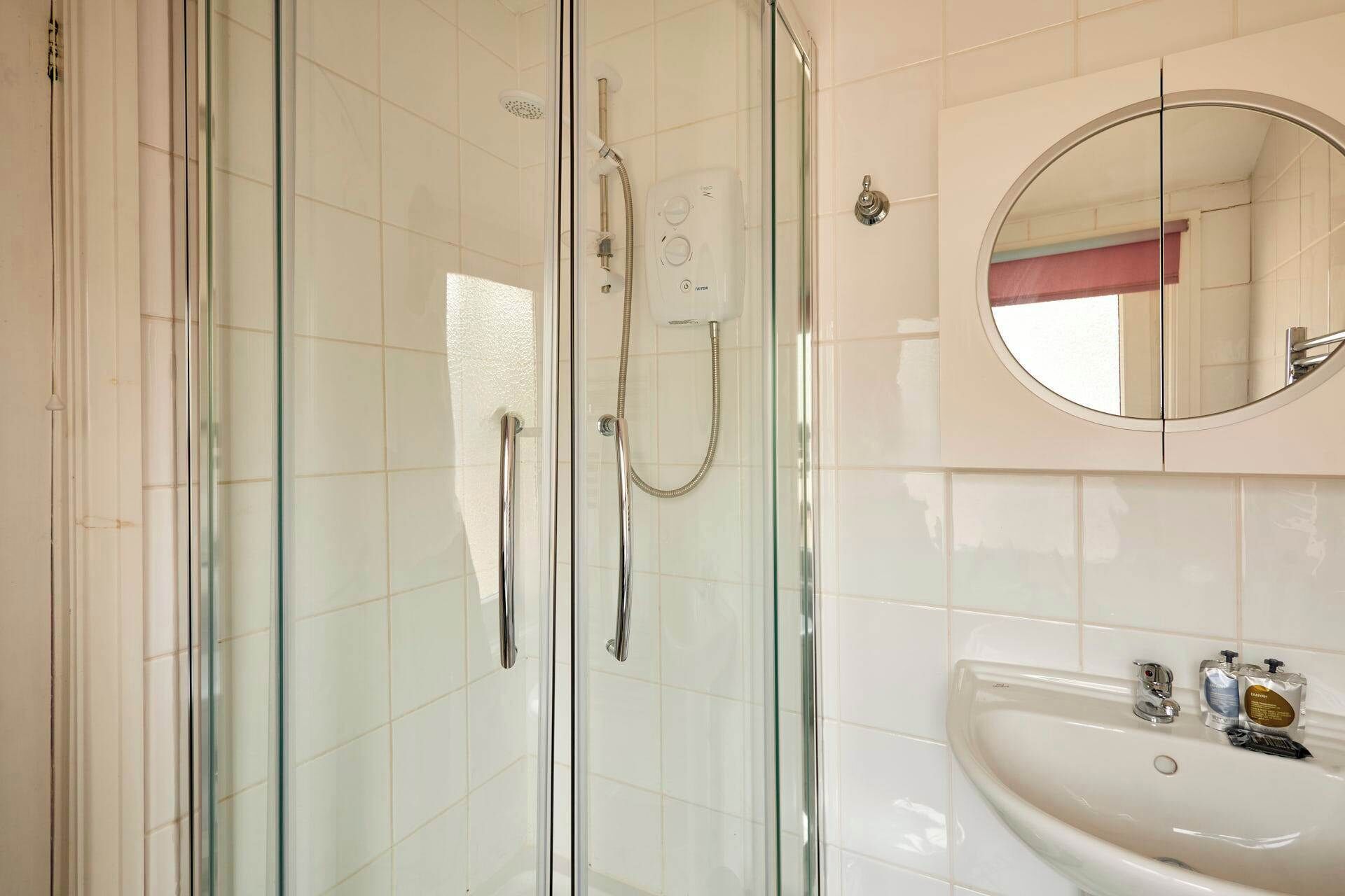 A bathroom with a glass-enclosed shower, a round mirror, and a white sink with toiletries.