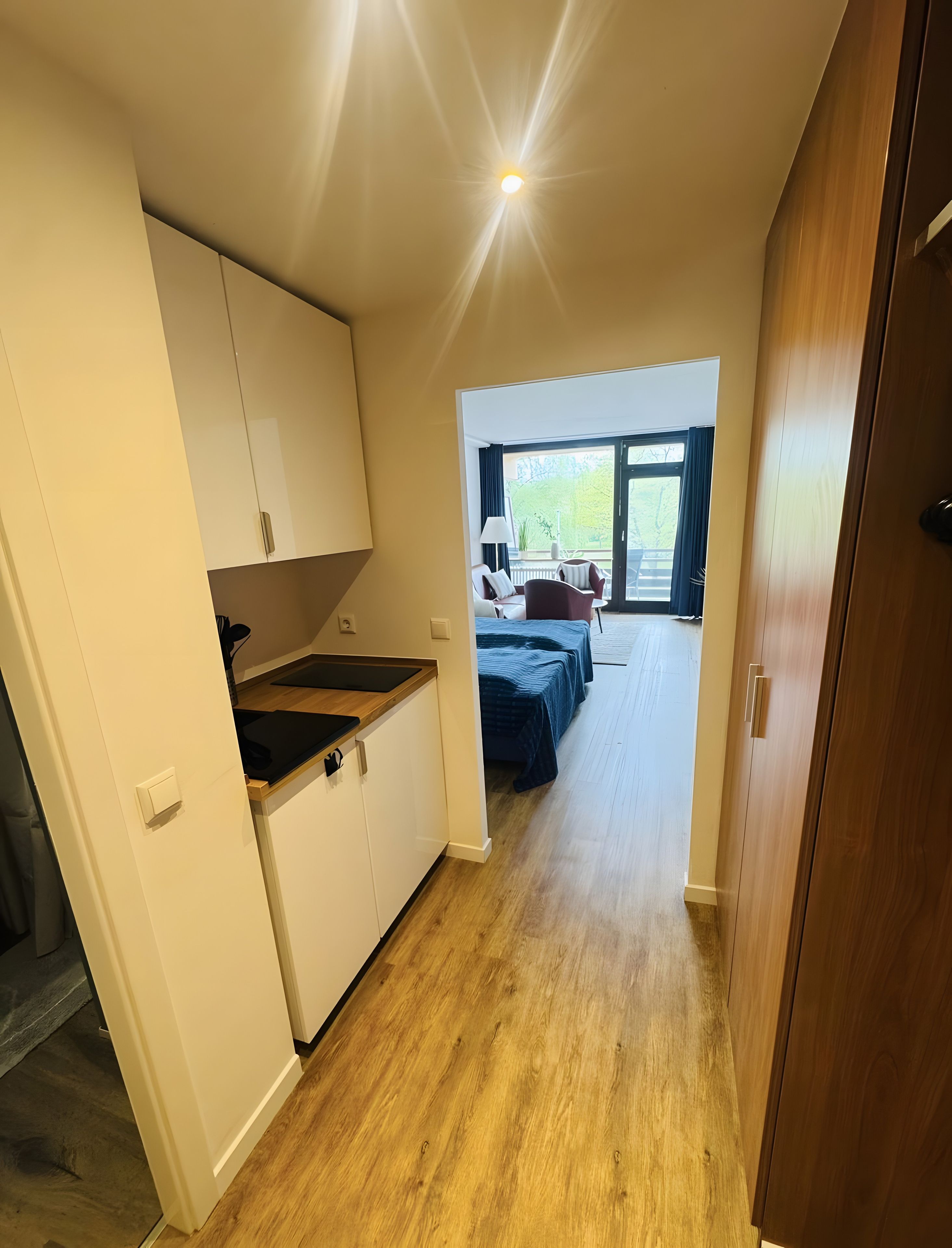 Ein enger Flur führt zu einem Schlafzimmer mit einem Bett, einem kleinen Tisch und einem Stuhl. Es gibt eine Küchenzeile mit Schränken und einer Arbeitsplatte.