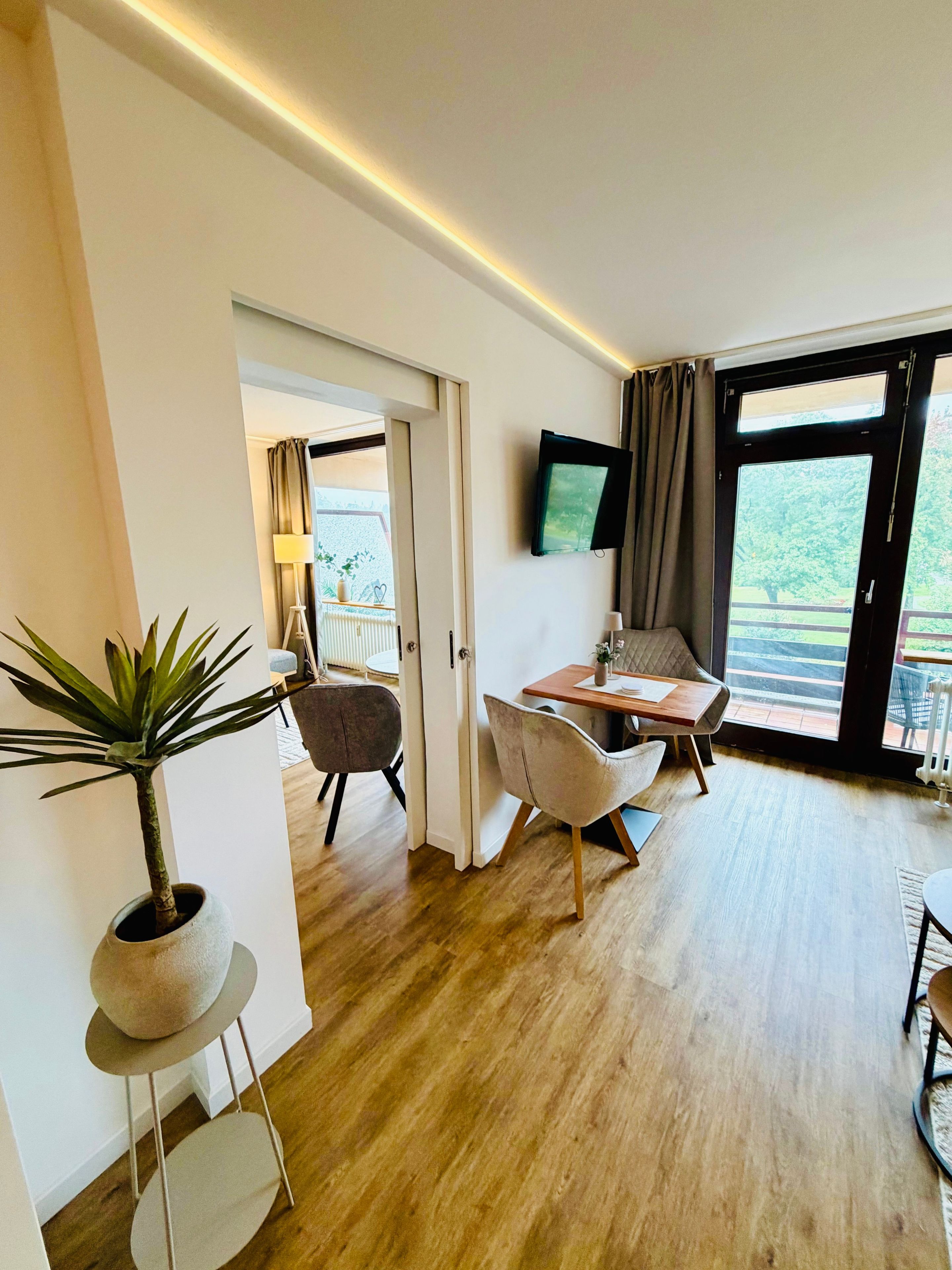 Ein modernes Zimmer mit Essbereich, einem an der Wand befestigten Fernseher und Blick ins Freie durch ein großes Fenster.