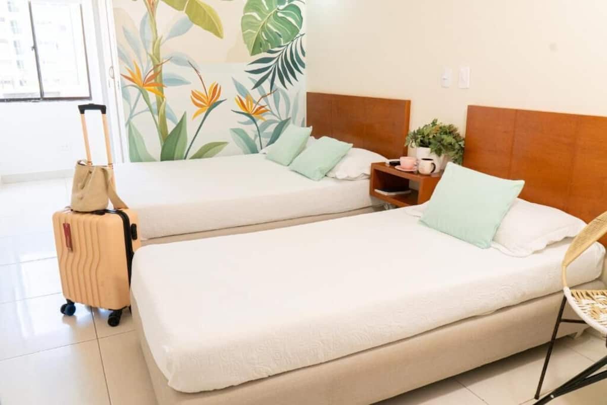 A hotel room with two beds, a suitcase, a chair, a nightstand, and a wall with a tropical plant mural.