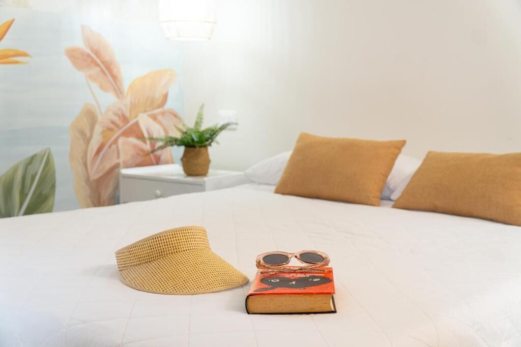 A neatly made bed with a book and sunglasses, a woven hat, and a potted plant on a bedside table.