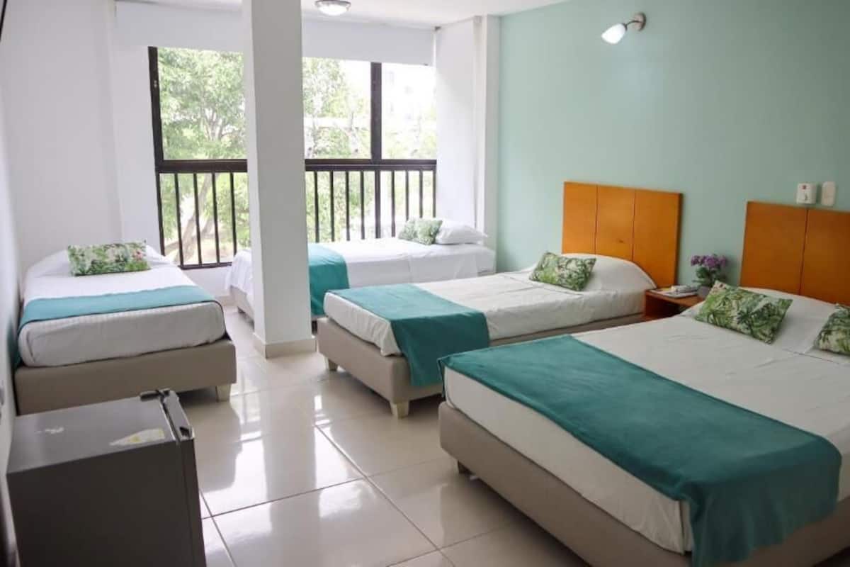 A hotel room with two single beds, a larger bed, a desk, and a balcony with a railing.