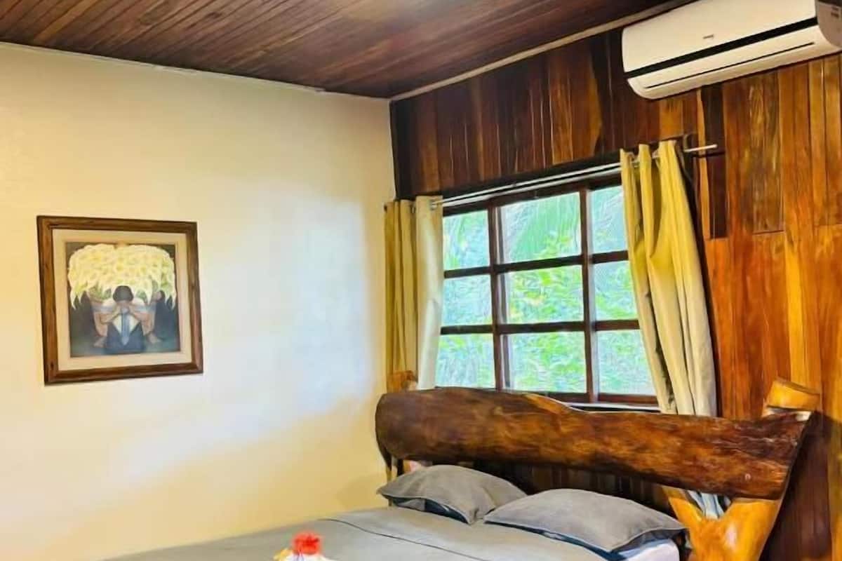 Una cama de madera con cabecera, una ventana con cortinas, un cuadro en la pared y un equipo de aire acondicionado.