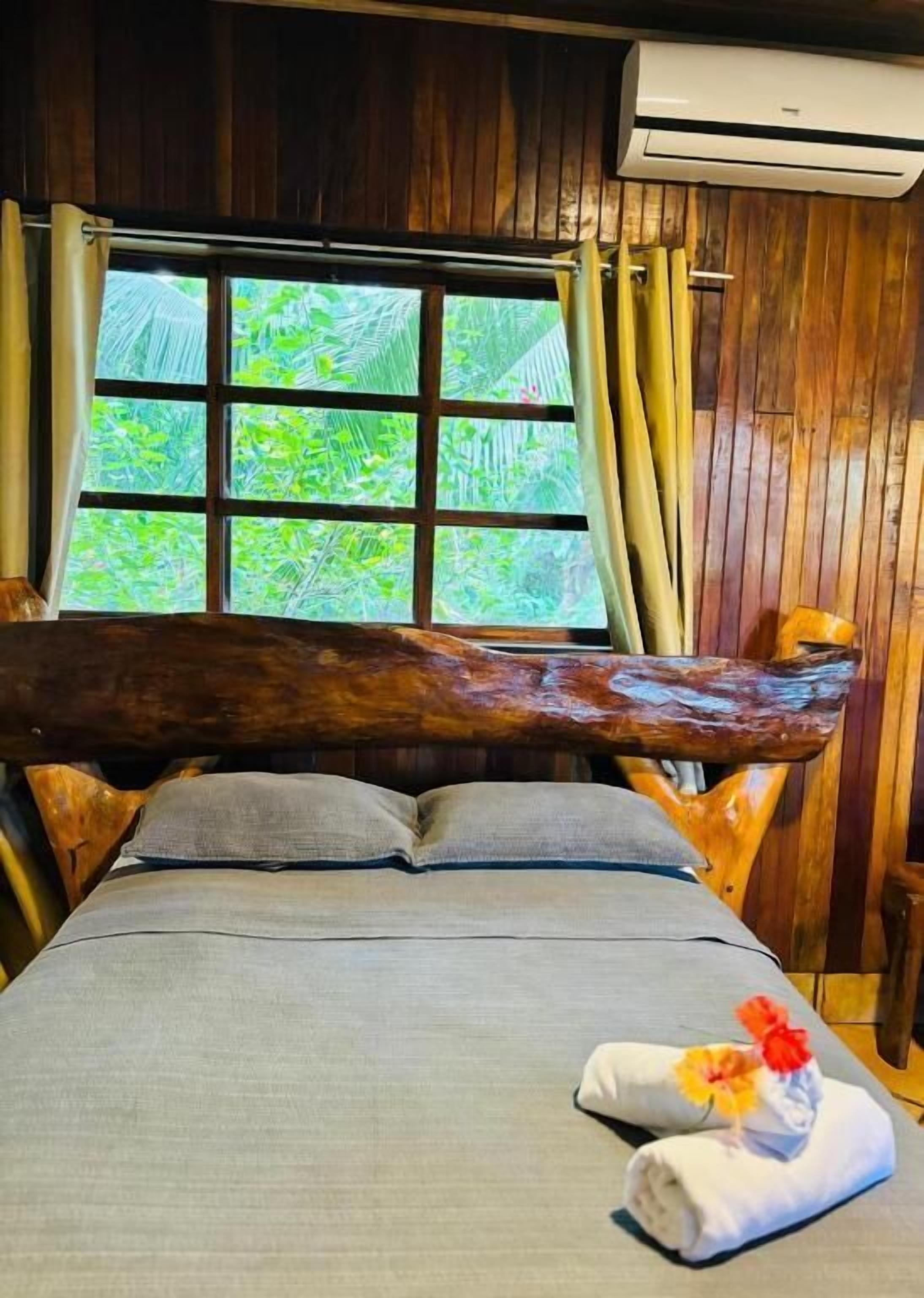 Un lit en bois avec une grande tête de lit en forme de tronc d’arbre, une fenêtre avec des rideaux et une vue sur la verdure à l’extérieur.