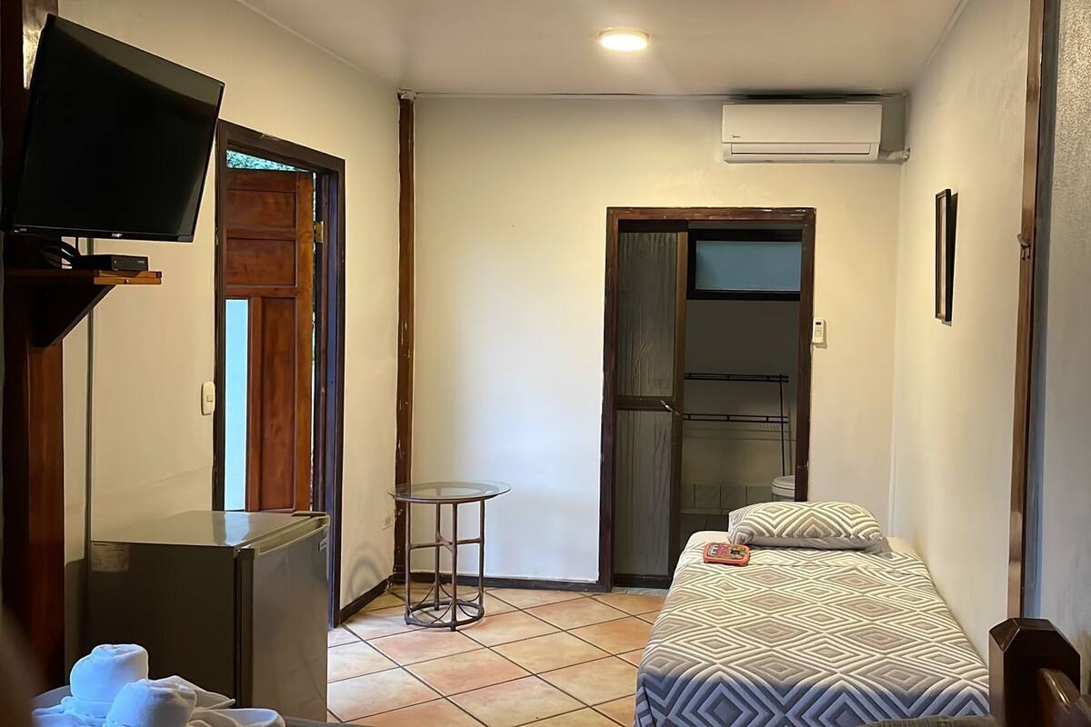 Una habitación pequeña y funcional con cama, televisión, una mesita y aire acondicionado.