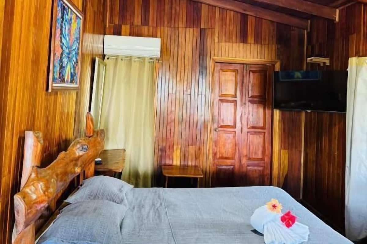 Una habitación de madera con una cama, cabecera de madera, una mesita, una puerta y un televisor.