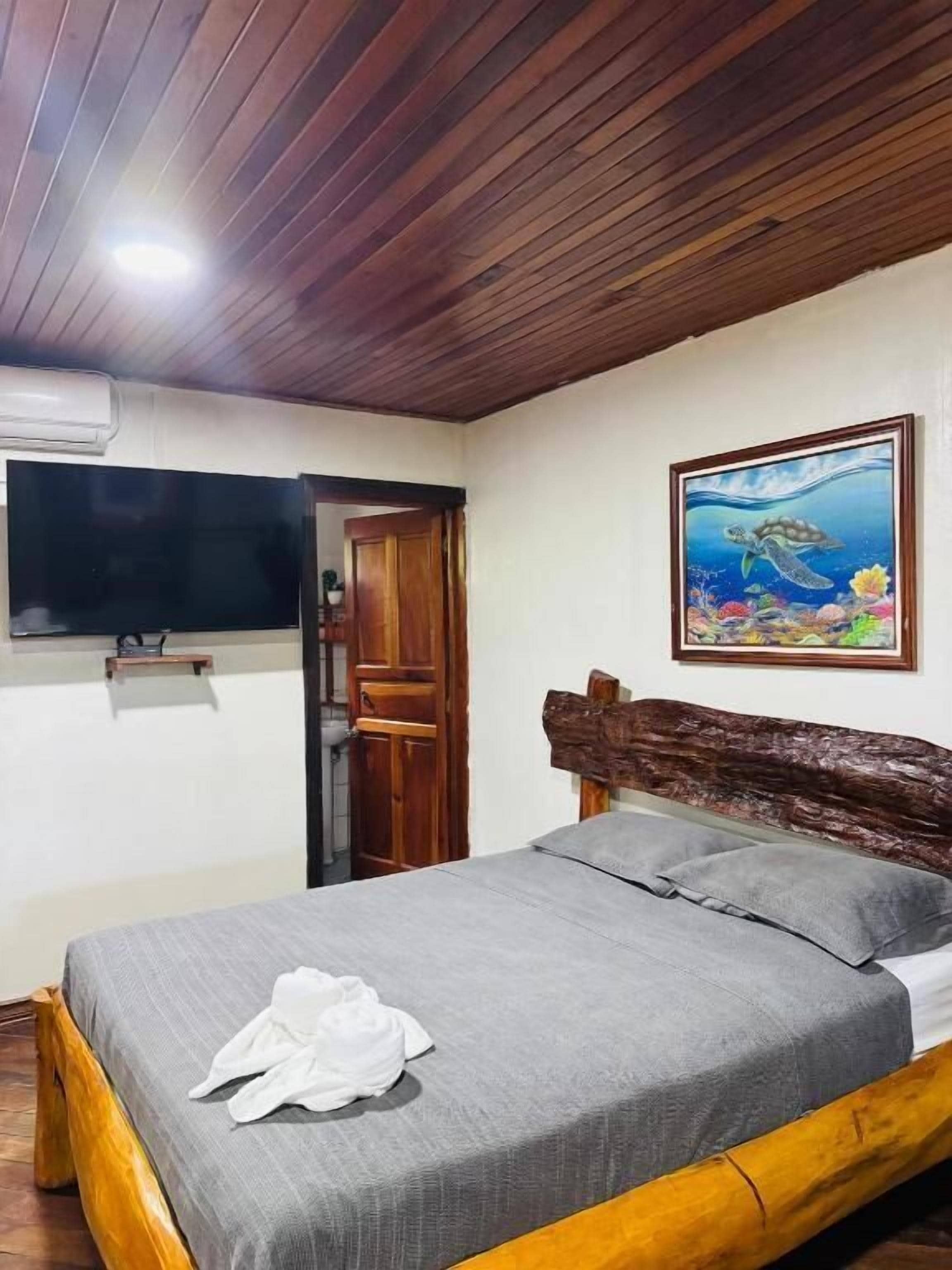 A bedroom with a wooden ceiling, a bed with grey bedding, a television mounted on the wall, and a painting of a turtle.