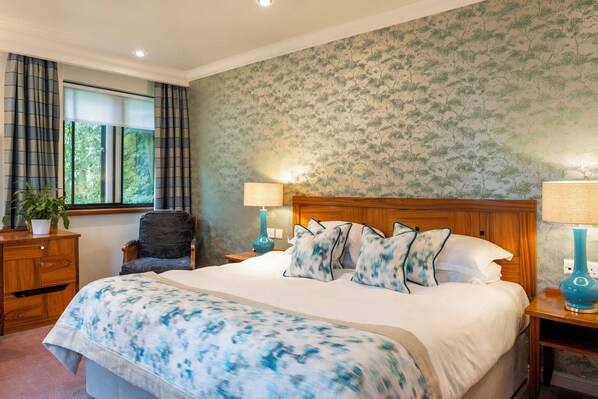 A bedroom with a large bed, wooden headboard, two bedside tables with lamps, a chair, a dresser, and a window with curtains.