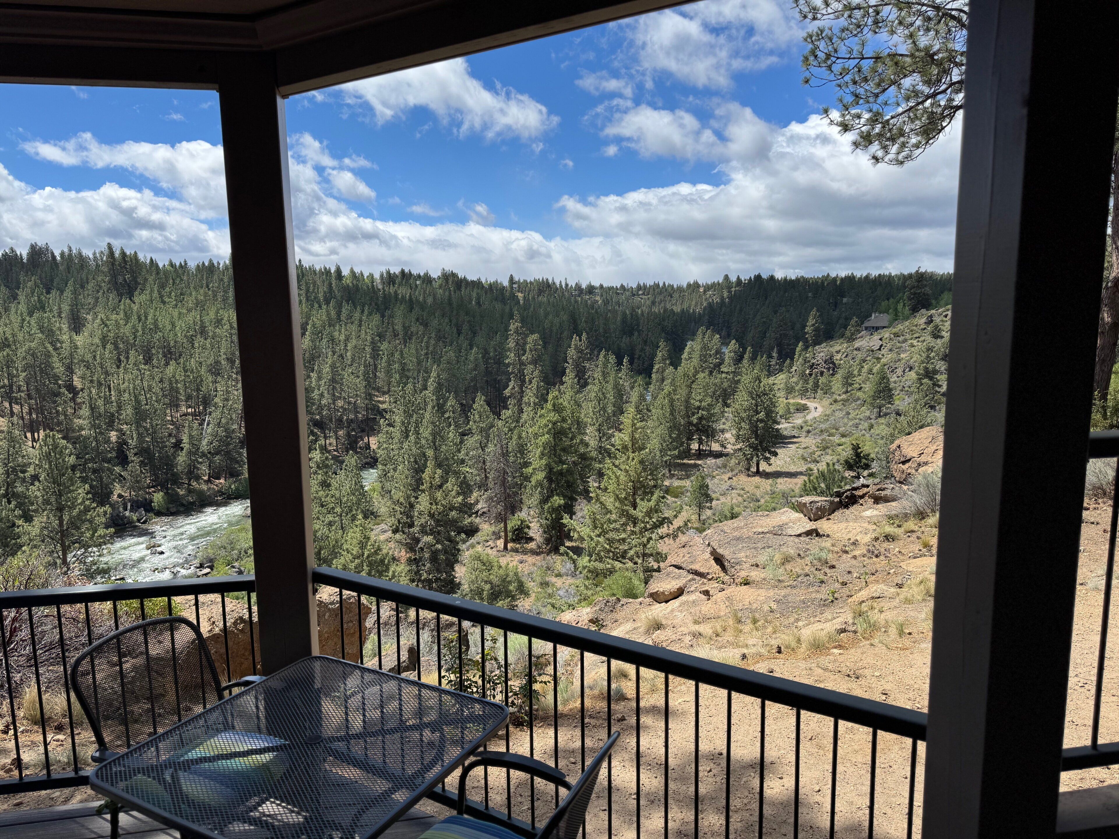 A balcony with a metal table and chairs, overlooking a forested valley with a river.