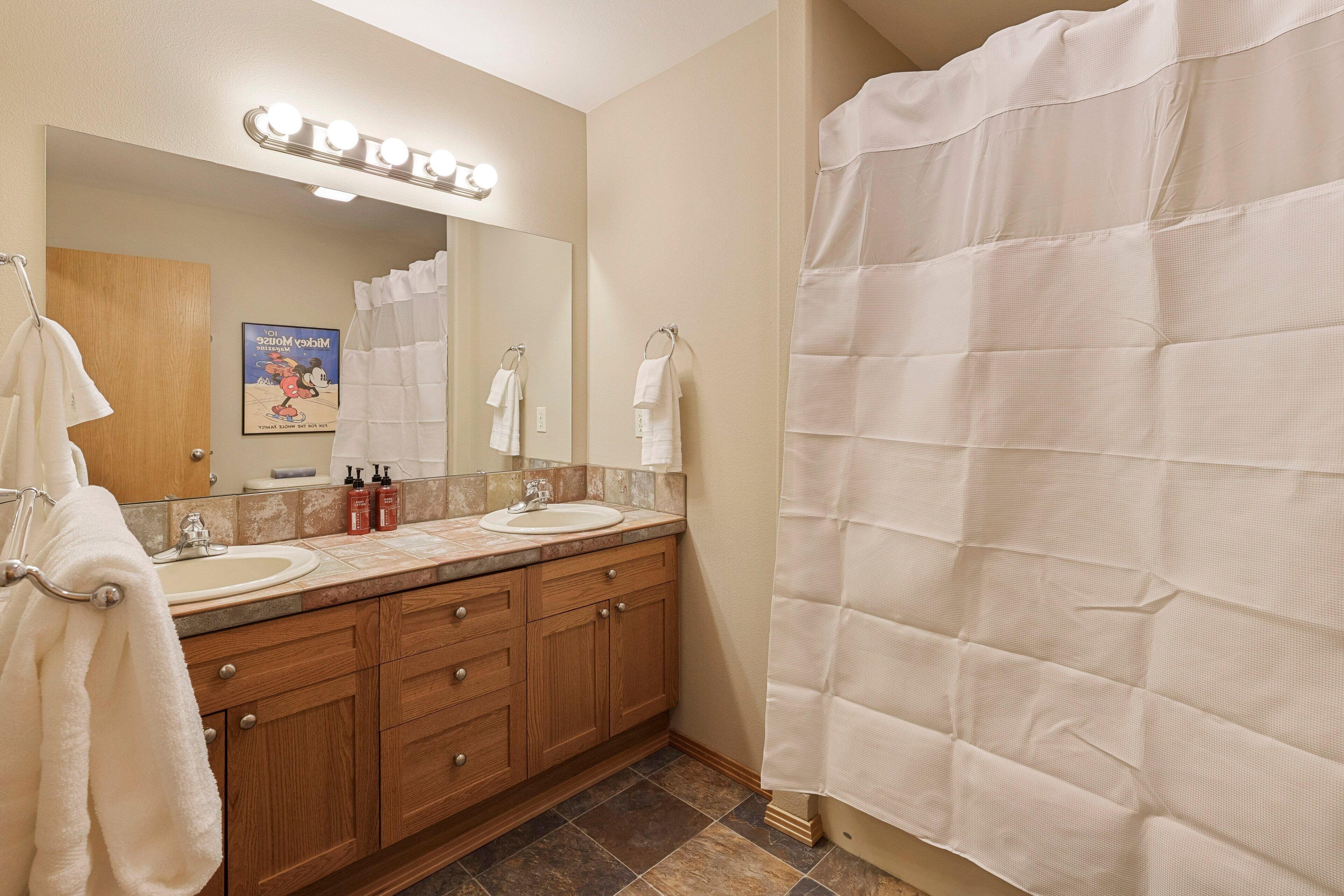 A bathroom with a double sink vanity, a mirror with lights, a shower curtain, and a Mickey Mouse poster on the wall.