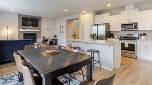 Uma cozinha moderna com armários brancos, uma ilha central, um espaço de refeições com mesa de madeira escura e uma sala de estar com sofá azul.