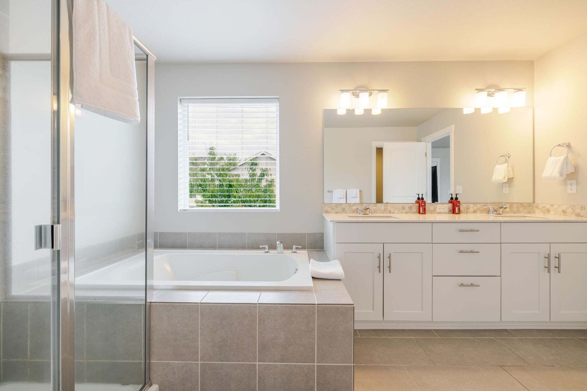 A modern bathroom with a glass shower enclosure, a large bathtub, a vanity with a mirror, and a window with blinds.