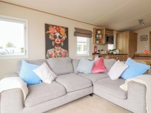 Ein Wohnzimmer mit grauem Ecksofa, einem Bild an der Wand und einer Küche im Hintergrund.