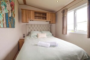 Ein ordentlich bezogenes Bett mit Kopfteil, ein kleines Holzschränkchen darüber und ein Fenster mit Vorhängen.