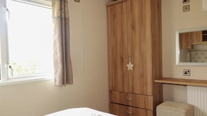 A bedroom with a bed, a wooden wardrobe, a small stool, a window with curtains, and a mirror above a wooden shelf. - Morwenna (Padstow)