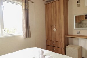 A bedroom with a bed, a wooden wardrobe, a small stool, a window with curtains, and a mirror above a wooden shelf.