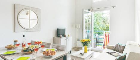 A modern, well-lit room with a dining table set for two, a sofa, a TV, and a balcony with outdoor seating.