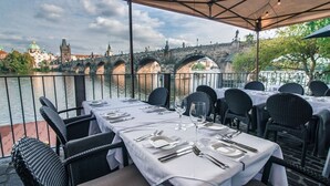 An outdoor dining area with tables set for two, overlooking a river with a bridge in the background.