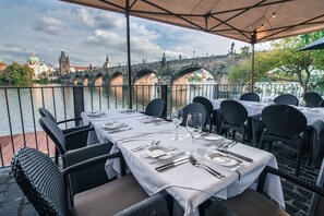 Un espacio de comedor al aire libre con mesas dispuestas para dos, con vistas a un río y un puente al fondo.