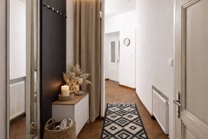 Ein Flur mit einer dunklen Tür, einem kleinen Regal mit Kerzen und getrockneten Blumen, einem gewebten Korb und einem gemusterten Teppich.