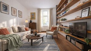 Een woonkamer met een bank, fauteuil, salontafel, televisie, boekenkast en kunstwerk aan de muur.