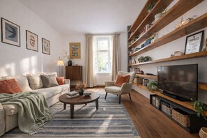Ein Wohnzimmer mit Sofa, Sesseln, Couchtisch, Fernseher, Bücherregal und Kunstwerken an der Wand.