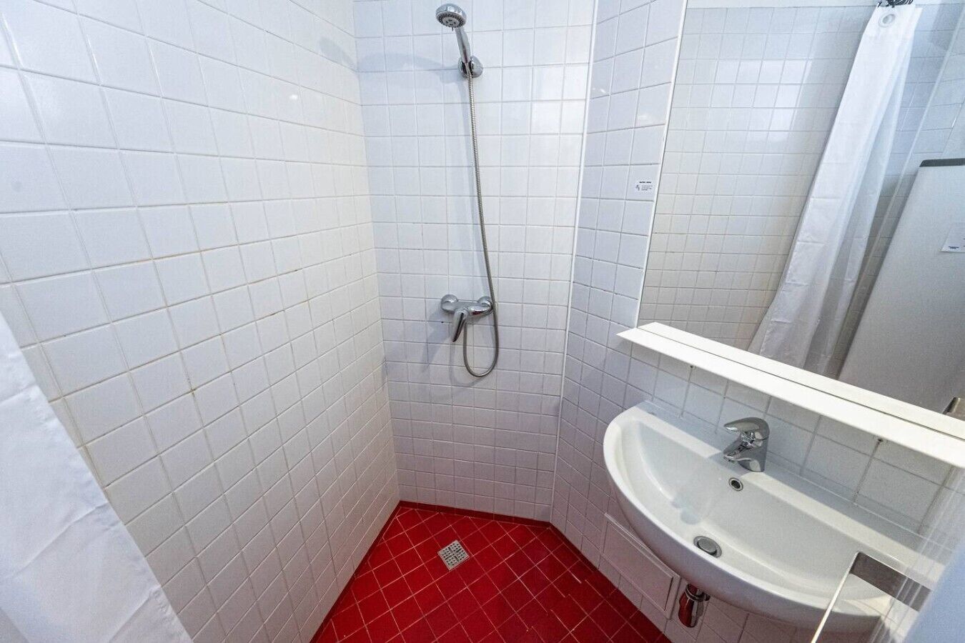 A bathroom with white tiled walls, a red floor, a white sink, and a shower.