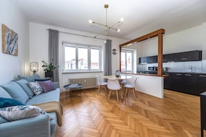 A modern living room with a sofa, dining table, and chairs, a kitchen area with dark cabinets, and large windows. - Urban Elegance Residence in Smíchov (Prague 5)