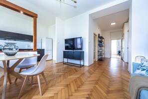 A modern apartment with a wooden herringbone floor, a dining area with a white table and chairs, a kitchen with dark cabinets, a TV on a black stand, and a living area with a sofa. - Urban Elegance Residence in Smíchov (Prague 5)