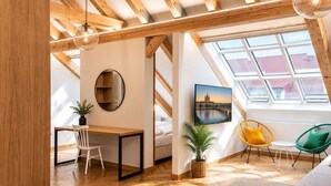 Una stanza con soffitto spiovente, travi in legno, una scrivania, una sedia, una pianta, un quadro e una finestra con vista.