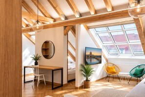 Ein Zimmer mit schrägen Wänden, Holzbalken, einem Schreibtisch, einem Stuhl, einer Pflanze, einem Gemälde und einem Fenster mit Aussicht.