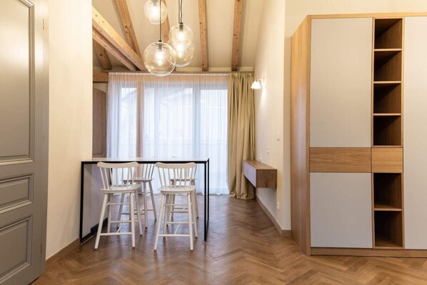 Ein Zimmer mit Holzboden, einem Esstisch mit Stühlen, einem großen Kleiderschrank und einem Fenster mit Vorhängen.