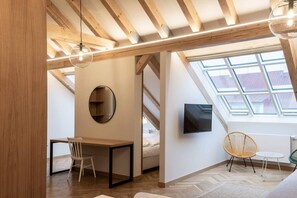 Ein Loft-Zimmer mit schrägverdeckter Decke, Holzbalken, Dachfenster, einem Bett, einem Fernseher, einem Schreibtisch und zwei Stühlen.