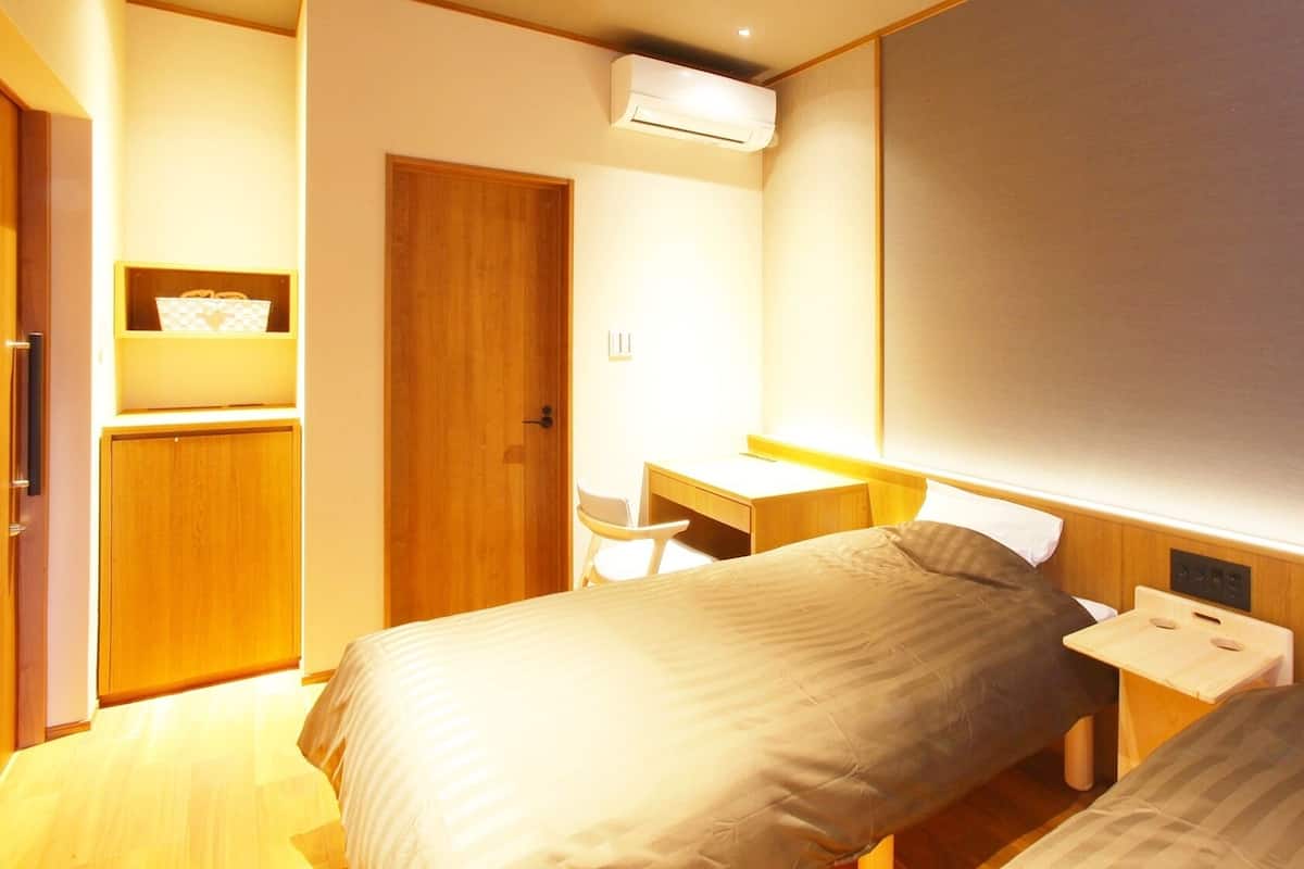 A hotel room with a bed, bedside table, desk, wardrobe, and an air conditioning unit.