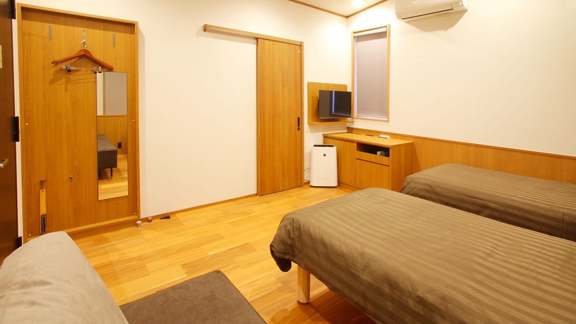 Quarto de hotel com duas camas, um guarda-roupa de madeira, uma escrivaninha com cadeira, um pequeno aquecedor e uma TV fixada na parede.