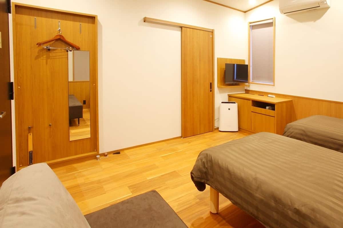 A hotel room with two beds, a wooden wardrobe, a desk with a chair, a small heater, and a TV mounted on the wall.