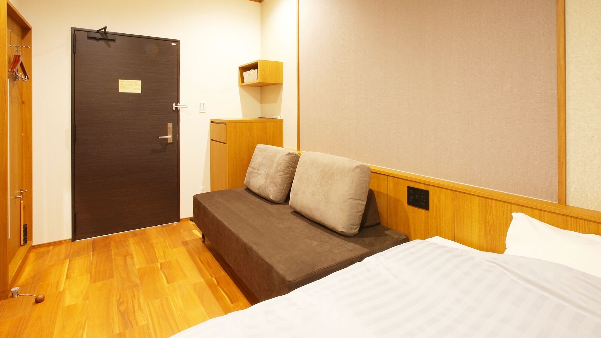 Quarto compacto com uma cama, um pequeno espaço para sentar, uma porta e uma prateleira na parede.
