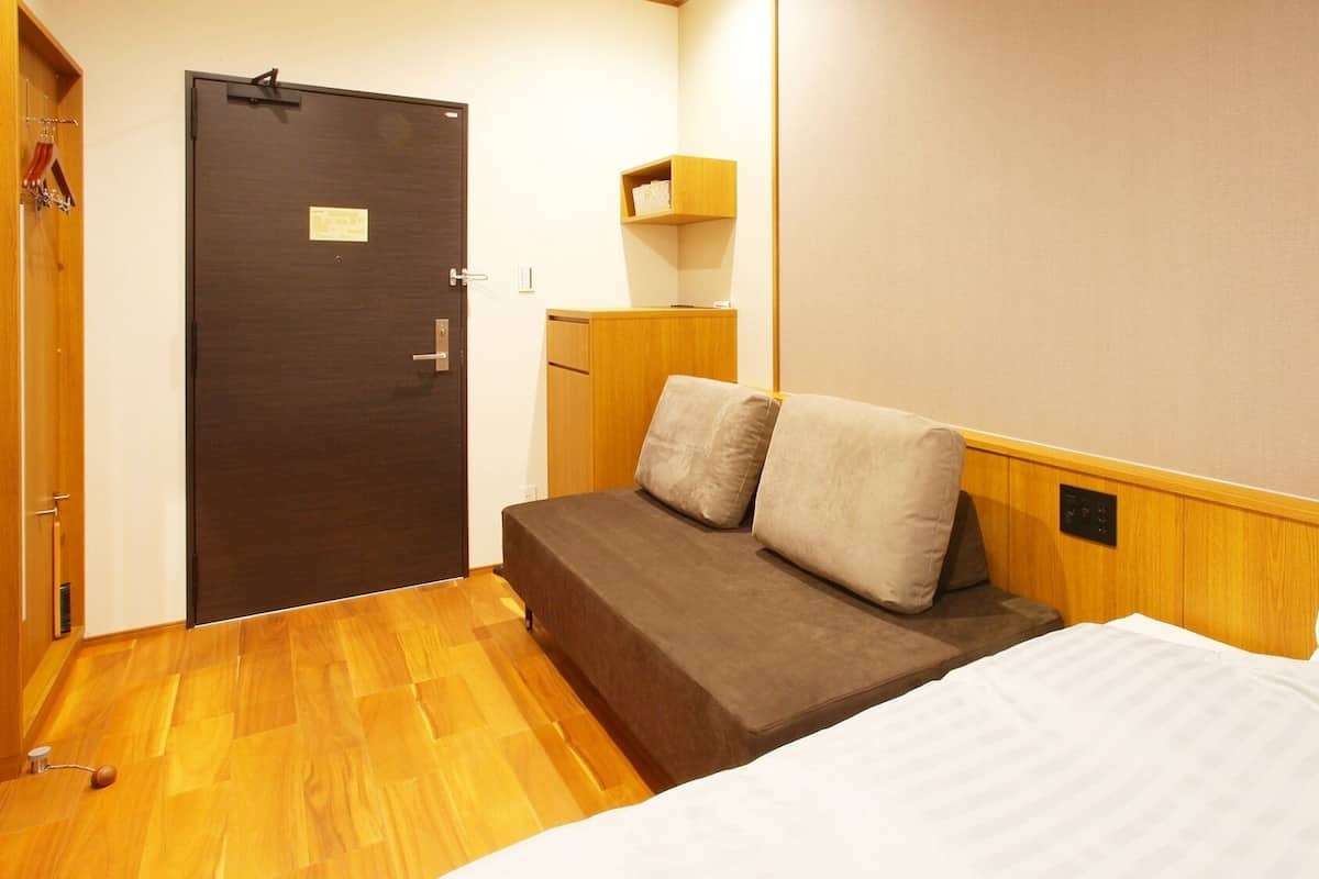 A compact room with a bed, a small seating area, a door, and a wall-mounted shelf.