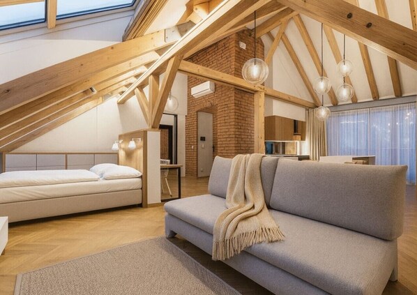 A loft-style room with a high ceiling, exposed wooden beams, a grey sofa, a white bed, and a brick wall. - Cozy Attic Loft with Terrace in Prime Location (Prague 2)