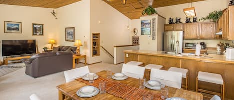 Un vaste espace de vie doté d’un plafond en bois, d’une table à manger pour six personnes, d’un canapé, d’un téléviseur et d’une cuisine avec des meubles et des appareils électroménagers en bois.