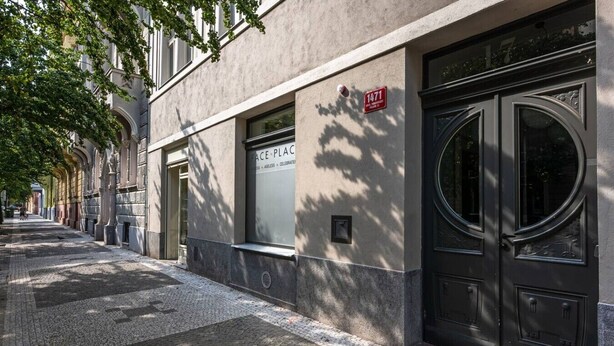 A building with a large window and a door, a street with cobblestone pavement, and a tree casting shadows. - Stylish Residence in Parkside Vinohrady (Prague 2)