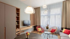 A modern living room with a round dining table, two red chairs, a grey sofa, a TV mounted on a wooden cabinet, and large windows with sheer curtains. - Stylish Residence in Parkside Vinohrady (Prague 2)