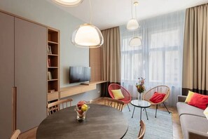 Ein modernes Wohnzimmer mit einem runden Esstisch, zwei roten Stühlen, einem grauen Sofa, einem an einem Holzschrank montierten Fernseher und großen Fenstern mit transparenten Vorhängen.