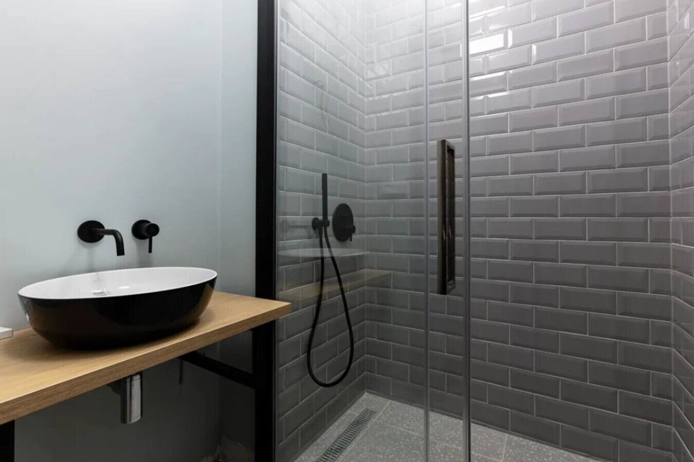 Une salle de bain moderne avec un lavabo sur pied noir, une douche à l’italienne en verre et des murs recouverts de carreaux gris.