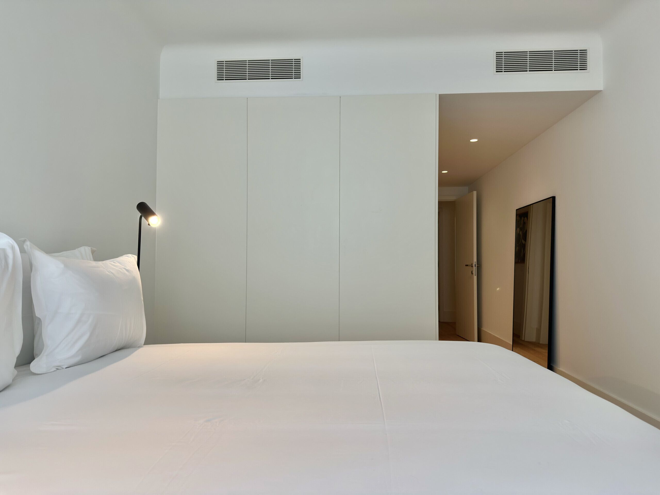 A bedroom with a bed, a bedside lamp, a wardrobe, and a door leading to another room.