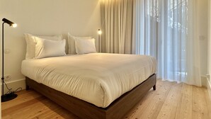 Un dormitorio con cama de madera, ropa de cama blanca, lámpara de noche y cortinas traslúcidas.