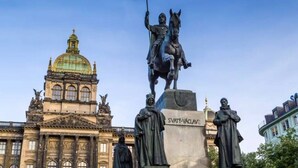 Une statue représentant une figure à cheval tenant un drapeau, un bâtiment surmonté d’un dôme et une autre statue devant.